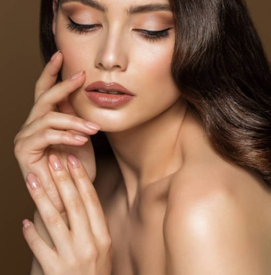 Beauty Model Face Make up with Beige Lipstick and Perfect Eyeliner on Closed Eyes. Woman Skin and Lips Care Cosmetics. Women Hands with Nude Nail Polish over Brown Beige Studio Background